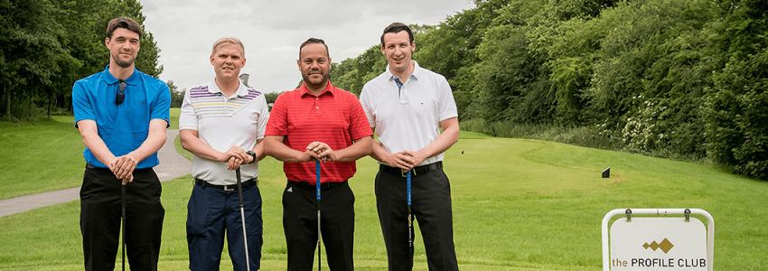 Four men stand on a lush green golf course, holding golf clubs and smiling at the camera.