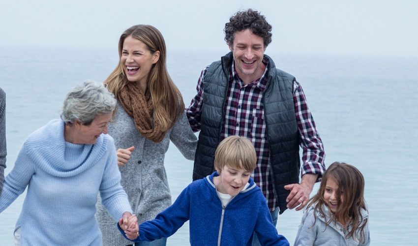 A family group of six, spanning three generations, is walking hand-in-hand along a sandy beach next to a calm sea. The weather appears overcast. The family consists of two older adults, a younger adult couple, and two young children.