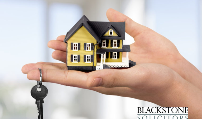 A hand gently holds a small, detailed model of a yellow and black two-story house in its palm. A single key on a keyring dangles below the hand.