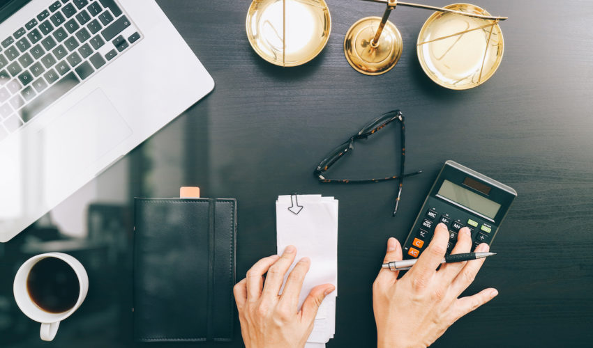 A top-down view of a dark wooden desk, shows a laptop, a brass scales of justice, eyeglasses, a calculator being used by a pair of hands, a notebook, and a cup of coffee.