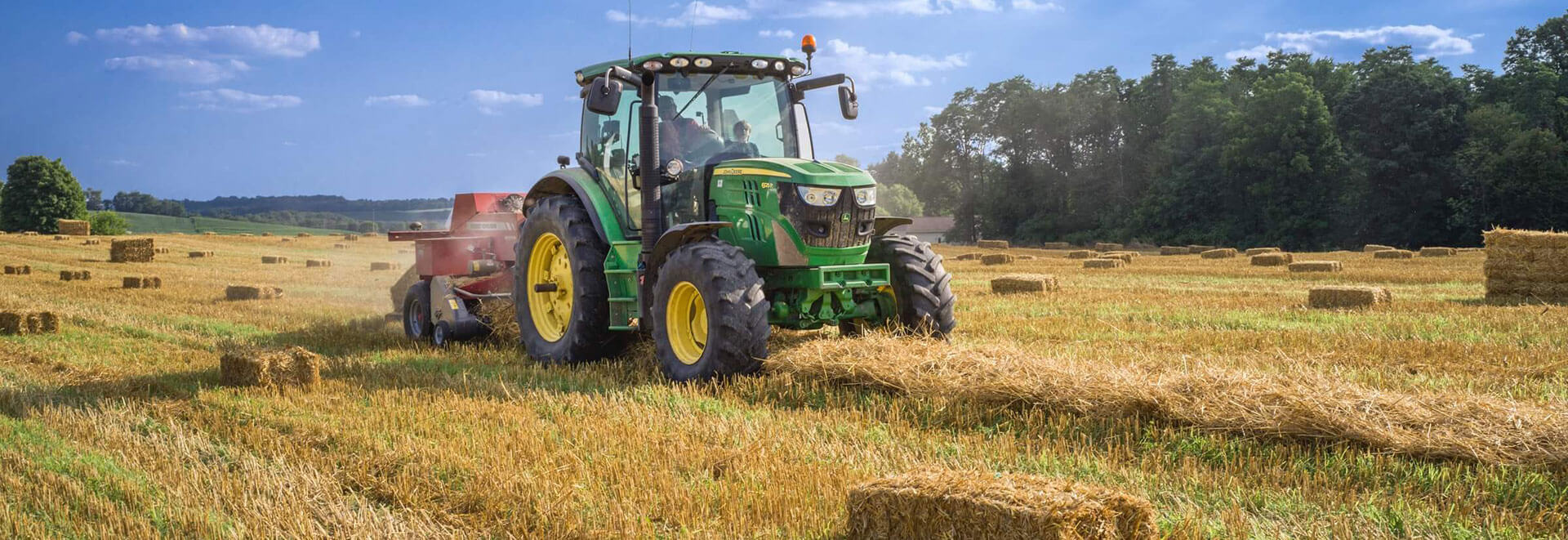 A green John Deere 6120M tractor with a baler works in a harvested field with scattered hay bales.