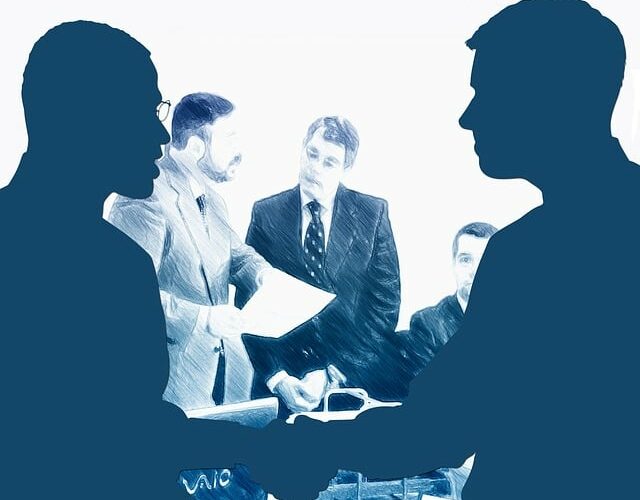 Businessmen shaking hands at a desk with colleagues in the background.