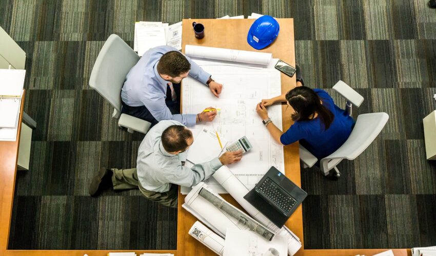 An overhead shot shows three people reviewing blueprints and using a calculator at a table with a blue hard hat and a laptop in an office.