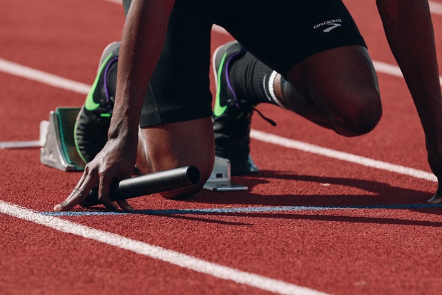 A runner in black shorts and green shoes is positioned at the starting line of a red track, holding a baton and using starting blocks.