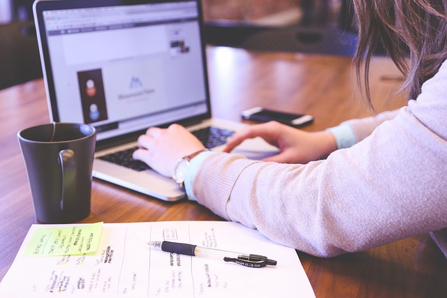 A person with long hair is working on a laptop at a wooden desk with a mug, papers, sticky notes, and a pen.