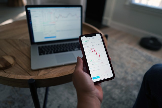 A person holds a smartphone displaying a stock market chart, with a laptop showing a similar chart in the background on a round wooden table.