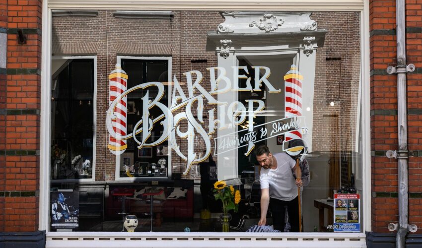 European-style barber shop window with lettering, poles, and a man inside.