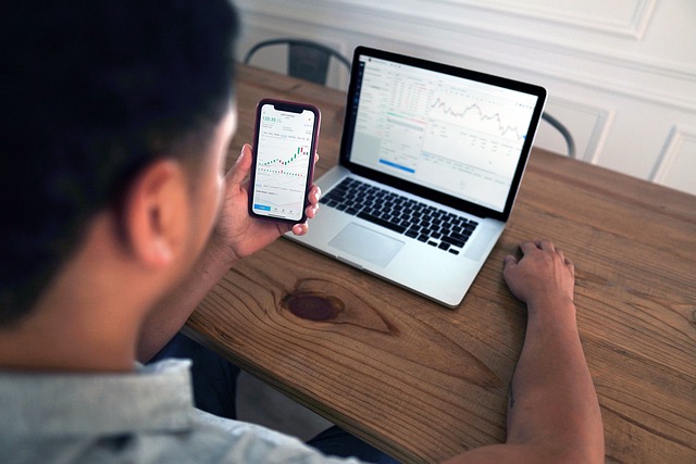 A person looks at stock market charts on a smartphone and a laptop on a wooden desk.