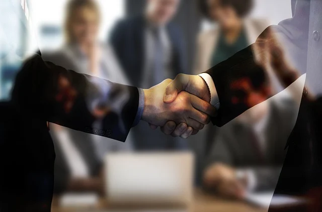 Two individuals in business attire are shaking hands in front of a blurred group of colleagues in an office setting.