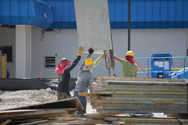 Three construction workers wearing hard hats and gloves guide a large concrete slab being lifted by machinery at a construction site.