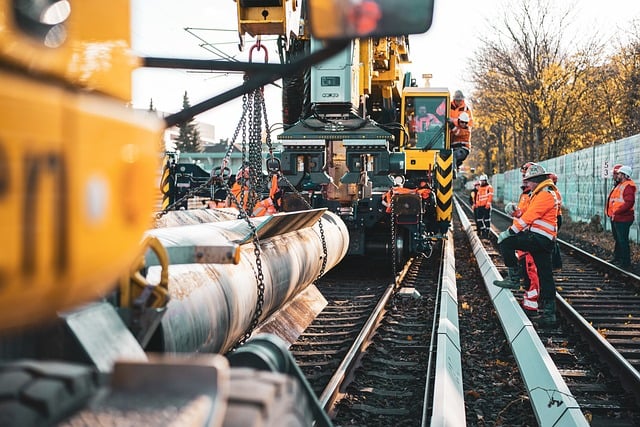 Construction workers in orange safety gear operate heavy machinery to lay a large pipe along a railway track.