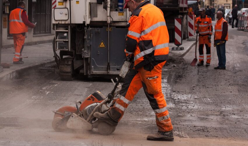 A construction worker in an orange high-visibility suit and a red cap uses a concrete saw on a city street, with other workers and equipment nearby.