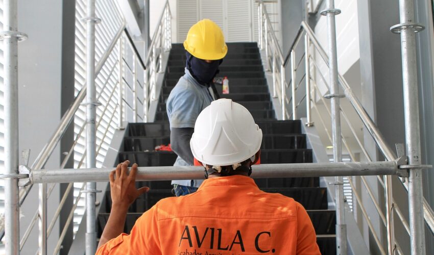 Two construction workers wearing hard hats, one white and one yellow, are on a staircase at a construction site.