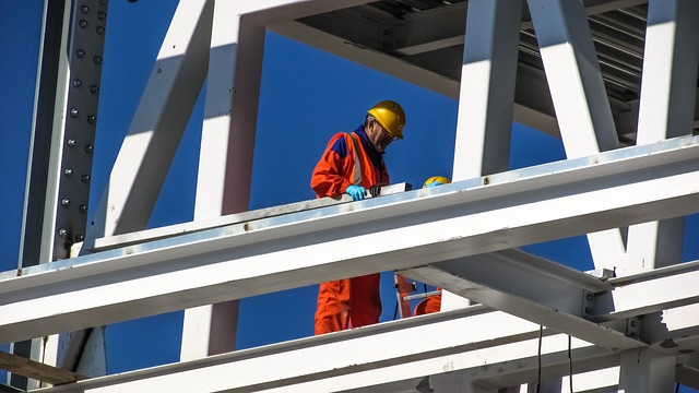 A construction worker in an orange jumpsuit, yellow hard hat, and blue gloves works on a white steel frame structure against a clear blue sky.