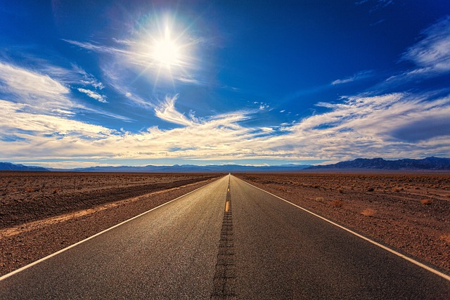 A long, straight road stretches into the distance under a bright sun and blue sky with scattered clouds.