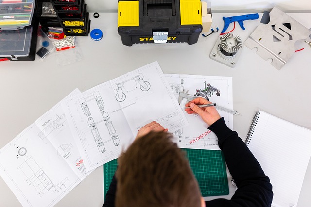 A person using a caliper to measure a technical drawing on a desk with tools and parts.