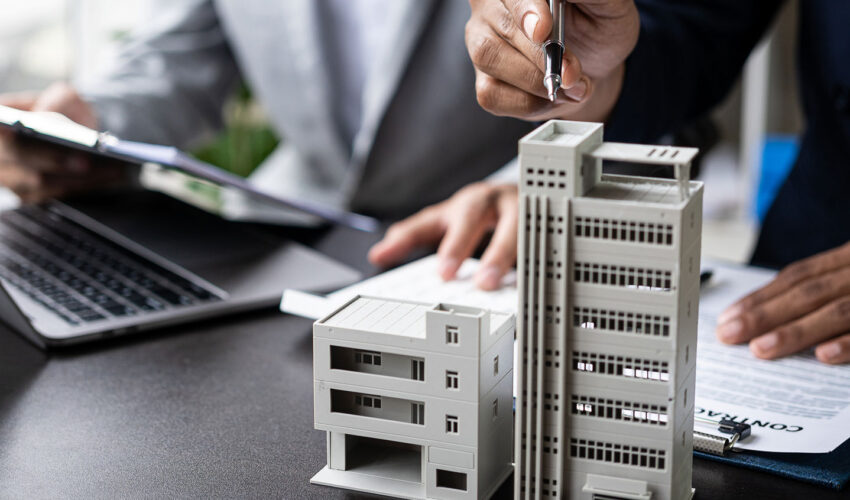 Two people discussing property finance with a miniature building model, documents, and a laptop on a desk.